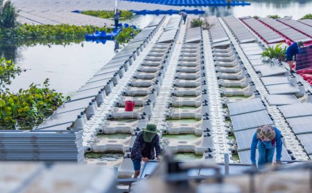 Foto de Un equipo de trabajadores maniobra cuidadosamente un panel solar en su lugar entre filas de paneles en una gran granja solar flotante, mostrando energía renovable en acción. - Imagen libre de derechos