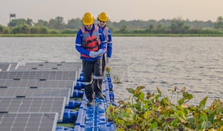 Foto de Dos ingenieros solares que usan equipo de protección y cascos inspeccionan visualmente las alineaciones, la estabilidad y la garantía de calidad de una gran variedad de paneles solares flotantes para cumplir con los requisitos estándar. - Imagen libre de derechos