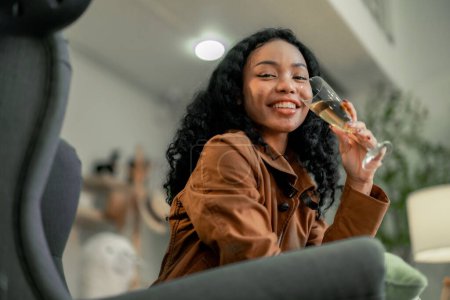 A young, black female startup designer relaxes with a glass of wine in an inviting workspace, reflecting on the day's accomplishments surrounded by her vivid creations.