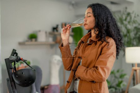 A young, black female startup designer relaxes with a glass of wine in an inviting workspace, reflecting on the day's accomplishments surrounded by her vivid creations.