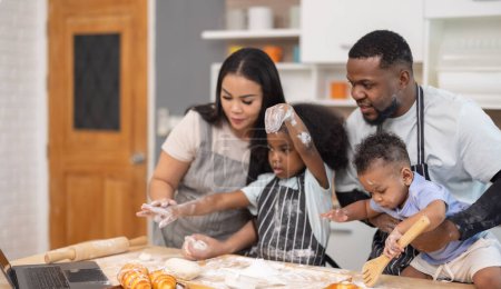 Téléchargez les photos : Une famille s'engage joyeusement dans la cuisson ensemble, en utilisant un ordinateur portable pour guider, entouré de farine et de petits pains. - en image libre de droit
