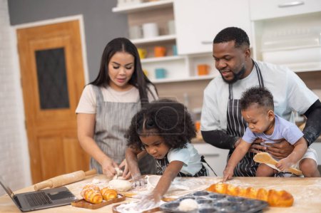 Téléchargez les photos : Une famille s'engage joyeusement dans la cuisson ensemble, en utilisant un ordinateur portable pour guider, entouré de farine et de petits pains. - en image libre de droit