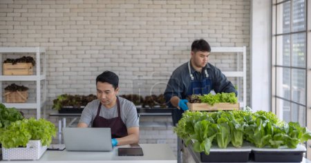Zwei Gemüseverkäufer, einer hantiert mit einem Laptop und der andere organisiert Grünzeug, demonstrieren eine effiziente Bestandsverwaltung innerhalb eines städtischen Marktes.