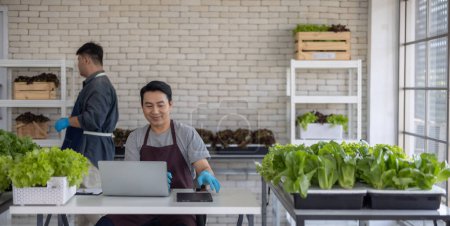 Two vegetable sellers, one handling a laptop and the other organizing leafy greens, demonstrate efficient inventory management inside an urban market.