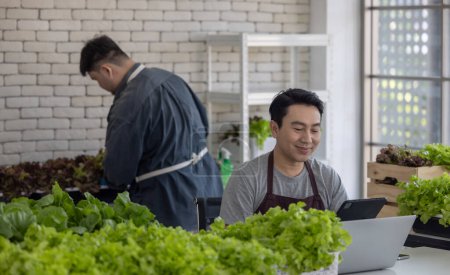 Deux vendeurs de légumes, l'un manipulant un ordinateur portable et l'autre organisant des légumes verts feuillus, font preuve d'une gestion efficace de l'inventaire dans un marché urbain.