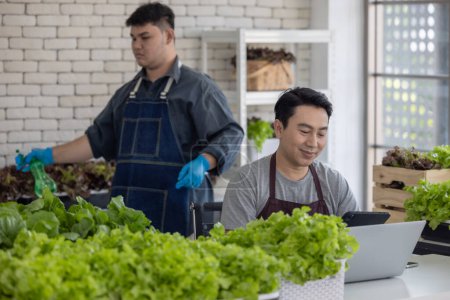 Two vegetable sellers, one handling a laptop and the other organizing leafy greens, demonstrate efficient inventory management inside an urban market.