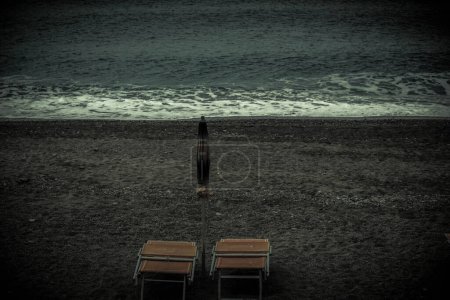 zwei Liegestühle und ein geschlossener Sonnenschirm an einem einsamen Strand am Ende des Sommers mit dunklem und düsterem Klima