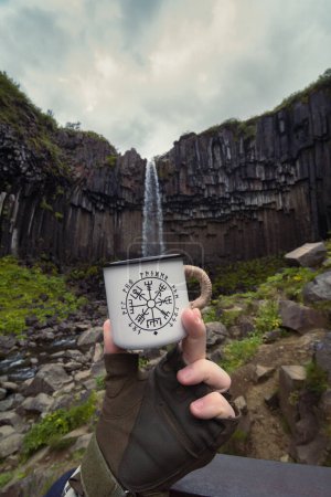 Photo for Close up man with mug against waterfall concept photo. Filling cup with water. First view hand photography with cliffs on background. High quality picture for wallpaper, travel blog, magazine, article - Royalty Free Image