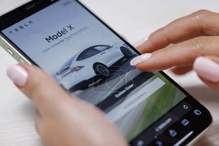 Photo for CALIFORNIA, USA - NOVEMBER 14, 2022: Close up of woman using smartphone shopping for a Tesla car browsing different models with the intent to purchase the vehicle online. Ordering Tesla model X Plaid. - Royalty Free Image