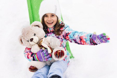 Téléchargez les photos : Portrait of child with toy teddy bear sits on a sled and looks at the winter snowy mountains.Winter vacantion. Christmas celebration and winter holidays. Winter fun and outdoor activities with kids. - en image libre de droit