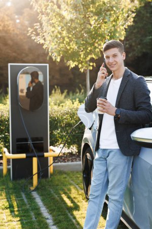 Photo for Portrait of handsome joyful 25-aged man which has mobile conversation while luxurious car charging battery on specially equipped charging station. - Royalty Free Image