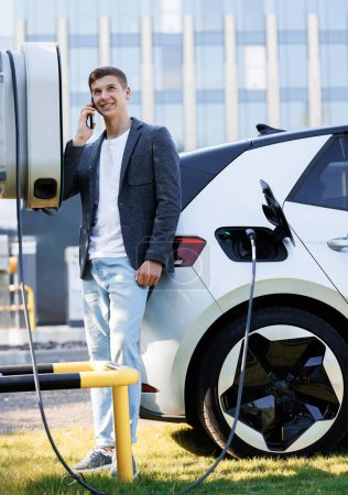 Photo for Portrait of handsome joyful 25-aged business man which has mobile conversation while luxurious electric car charging battery on specially equipped charging station. Charging electric car. - Royalty Free Image