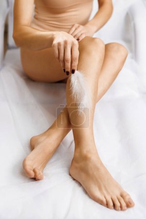 Primer plano de las piernas femeninas con piel lisa y pluma de avestruz suave. Conceptos de cuidado de la piel y tratamiento de depilación. Mujer toca sus piernas con pluma blanca.