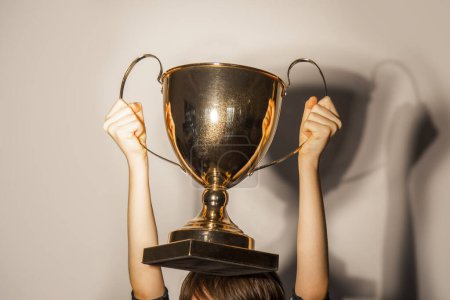 Photo for Hands holding a golden trophy close-up against white wall with shadows. Winning success achieve the goal, leadership, award concept - Royalty Free Image