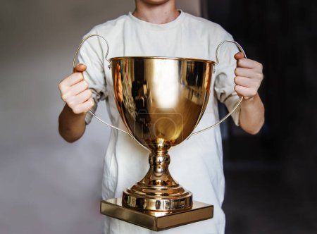 A boy in a white T-shirt holds a golden trophy close-up. Winning success achieve the goal, leadership, award concept