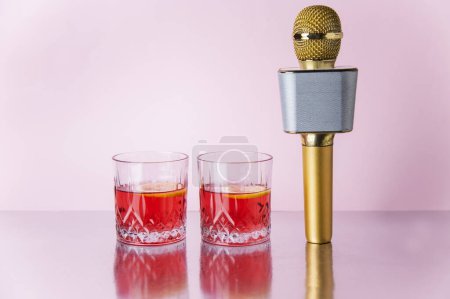 A vertically standing microphone and two glasses of pink cocktail on a reflection pink background isolated with copy space. Karaoke concept 