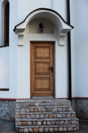 Foto de A light brown wooden door with a black lock, above the door is a lantern, the building is white, the lower part is decorated with stones and stairs - Imagen libre de derechos