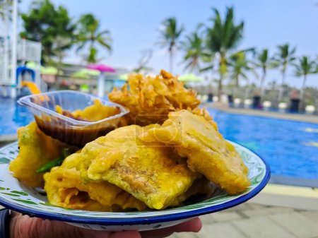 Téléchargez les photos : Profitant de beignets indonésiens au bord de la piscine. Bakwan Sayur et Tempe Mendoan dans une assiette au piment vert et sauce Petis, piscine floue en arrière-plan - en image libre de droit