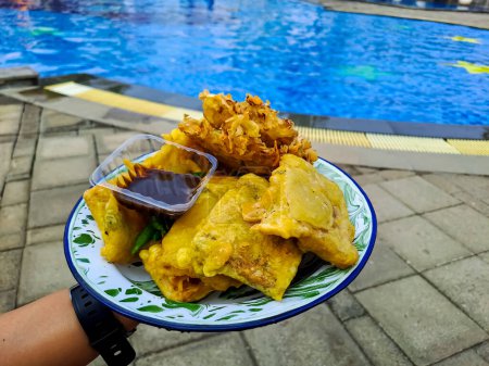Téléchargez les photos : Profitant de beignets indonésiens au bord de la piscine. Bakwan Sayur et Tempe Mendoan dans une assiette au piment vert et sauce Petis, piscine floue en arrière-plan - en image libre de droit