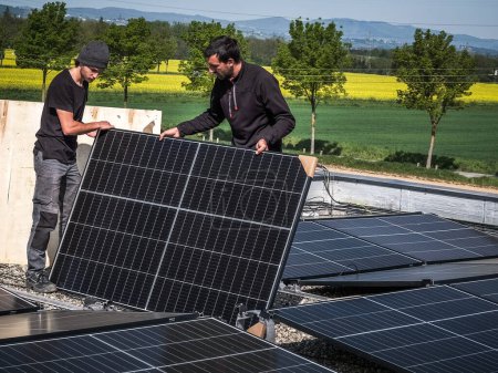 Foto de Male team engineers installing stand-alone solar photovoltaic panel system. Electricians mounting blue solar module on roof of modern house. Alternative energy concept - Imagen libre de derechos