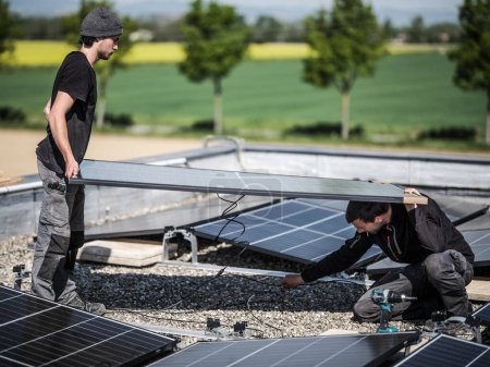 Foto de Male team engineers installing stand-alone solar photovoltaic panel system. Electricians mounting blue solar module on roof of company. Alternative energy concept - Imagen libre de derechos
