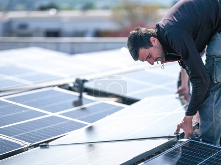 Foto de Male team engineers installing stand-alone solar photovoltaic panel system. Electricians mounting blue solar module on roof of company roof. Alternative energy concept - Imagen libre de derechos