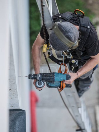 Foto de An electrical engineer of the team installs the electrical cables for the autonomous photovoltaic solar panel system. It is installed on a scale and uses a meter and a pencil for position measurements and a drill. For his safety he has a harness conn - Imagen libre de derechos