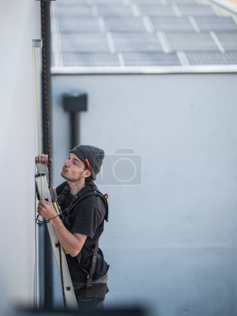 Foto de An electrical engineer of the team installs the electrical cables for the autonomous photovoltaic solar panel system. It is installed on a scale and uses a meter and a pencil for position measurements and a drill. For its safety it has a harness conn - Imagen libre de derechos