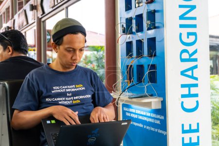 Téléchargez les photos : Cengkareng, 26 novembre 2023 : Le personnel informatique travaille sur un ordinateur portable par une station de charge de batterie gratuite dans le terminal de l'aéroport de Soekarno Hatta - en image libre de droit
