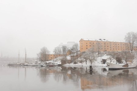 Scandinavian style historic yellow stone buildings with white snow on the ground on Skeppsholmen, Stockholm, Sweden by the water with a small boat marina during heavy snowfall in winter