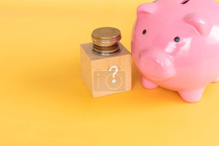 Foto de Ahorro de dinero; Un bloque de madera con un signo de interrogación, una alcancía rosa, y monedas. - Imagen libre de derechos