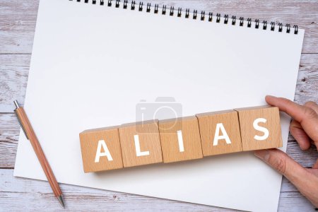 Holzblöcke mit Konzepttext "ALIAS", einem Stift, einem Notizbuch und einer Hand.