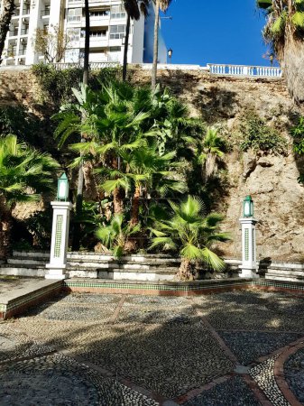 Amidst the beauty is a public garden decorated with carefully placed rocks, sunlight reflecting on it like a silent canvas, tiled columns, and their mosaic patterns with palm trees in the background.
