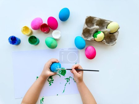 Childrens hands paint Easter eggs with brush surrounded by colorful eggs and jars of paints on white table, creative holiday activity. High quality photo