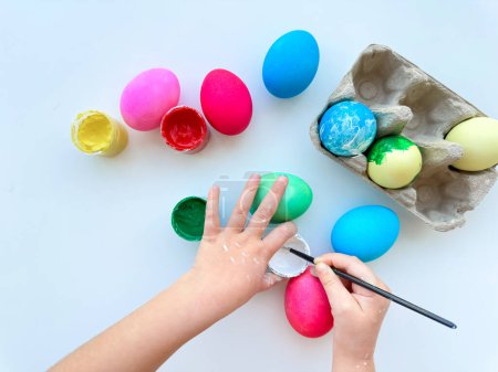 Kinderhände bemalen Ostereier mit Pinsel, umgeben von bunten Eiern und Farbgläsern auf weißem Tisch, kreative Ferienaktivitäten. Hochwertiges Foto