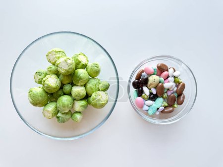 Brotes frescos de Bruselas en un tazón de vidrio junto a un tazón de dulces de colores sobre fondo blanco. Concepto de comida saludable versus indulgente. Foto de alta calidad