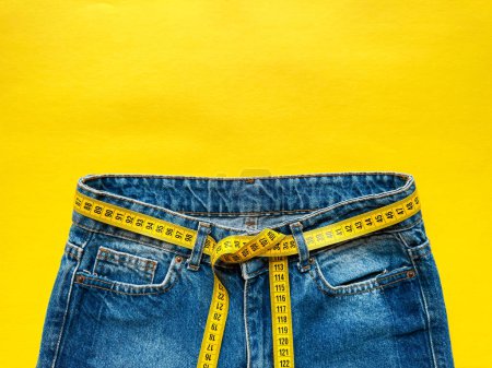 Blaue Jeans mit Maßband als Gürtel auf hellgelbem Hintergrund mit Kopierraum. Darstellung von Gewichtsverlust, Ernährung und gesundem Lebensstil. Ansicht von oben. Hochwertiges Foto