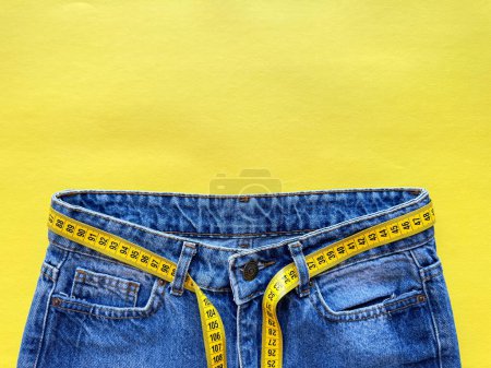 Blaue Jeans mit Maßband als Gürtel auf hellgelbem Hintergrund mit Kopierraum. Darstellung von Gewichtsverlust, Ernährung und gesundem Lebensstil. Ansicht von oben. Hochwertiges Foto
