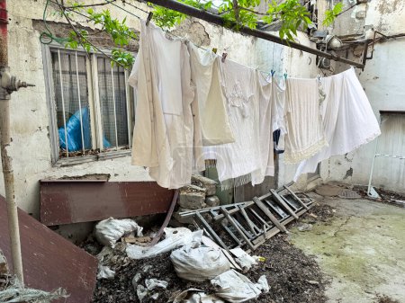 Laundry hangs on line in dilapidated city yard with overgrown leaves, broken furniture and trash, depicting neglect and everyday life and poverty in run down neighborhood. High quality photo