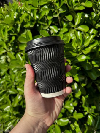 Mano sosteniendo taza de café de cartón corrugado negro con tapa contra fondo de frondoso arbusto verde exuberante. Estilo de vida ecológico urbano. Espacio en blanco para pegatina o etiqueta. Foto de alta calidad