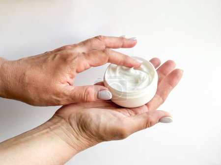 Close up of middle age woman hands holding container of cream and dipping finger on white background. Skincare and moisturizing concept. For healthcare, wellness, and self care poster. High quality