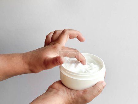 Close up of childs hands holding container of hand cream and dipping finger on white background. Skincare and moisturizing concept. Design for healthcare, wellness, and child care poster. High quality
