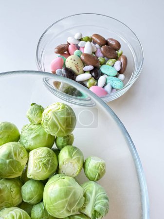 Brussels sprouts in clear glass bowl above assorted candy in another bowl on white surface. Healthy eating concept. High quality photo
