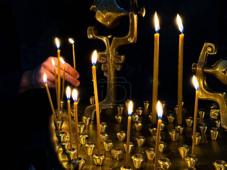 Hand lighting candles in dark room with golden candle holder. Religious or spiritual ceremony with candlelight. Lighting a candle in an Orthodox or Catholic church. High quality photo