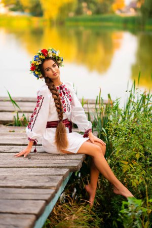 Photo for Beautiful young girl with extra long braided hair in traditional ukrainian national clothes in a wreath of colorful flowers sitting on nature near river. - Royalty Free Image