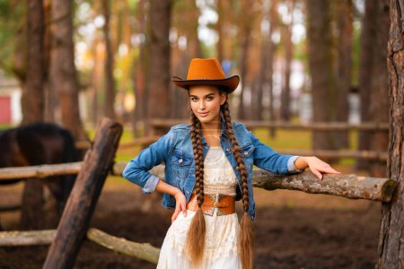 Portrait d'une belle cow-girl aux cheveux tressés extra longs et robe vintage à la campagne, posant dans la forêt.