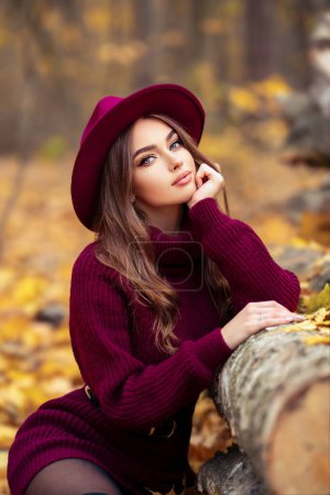 Foto de Hermosa chica en acogedor vestido de punto borgoña y sombrero sentado en la naturaleza con fondo de otoño. Retrato de una modelo bonita. - Imagen libre de derechos