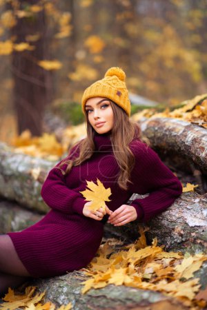 Photo for Beautiful girl in cosy knitted burgundy dress and hat sitting on nature with autumn background. Pretty model with leaf. - Royalty Free Image