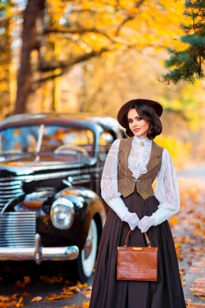 Schöne Frau in Vintage-Kleid, Spitzenbluse und Hut mit Schleier steht in der Nähe von retro braunen Auto auf herbstlichem Hintergrund. Elegante Dame in Handschuhen posiert auf der Natur.Warmes Kunstwerk.
