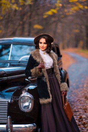 Téléchargez les photos : Belle femelle en robe vintage, chemisier en dentelle et chapeau avec voile debout près de voiture brun rétro sur fond d'automne. Élégante dame en gants posant sur la nature.Travail d'art chaud. - en image libre de droit
